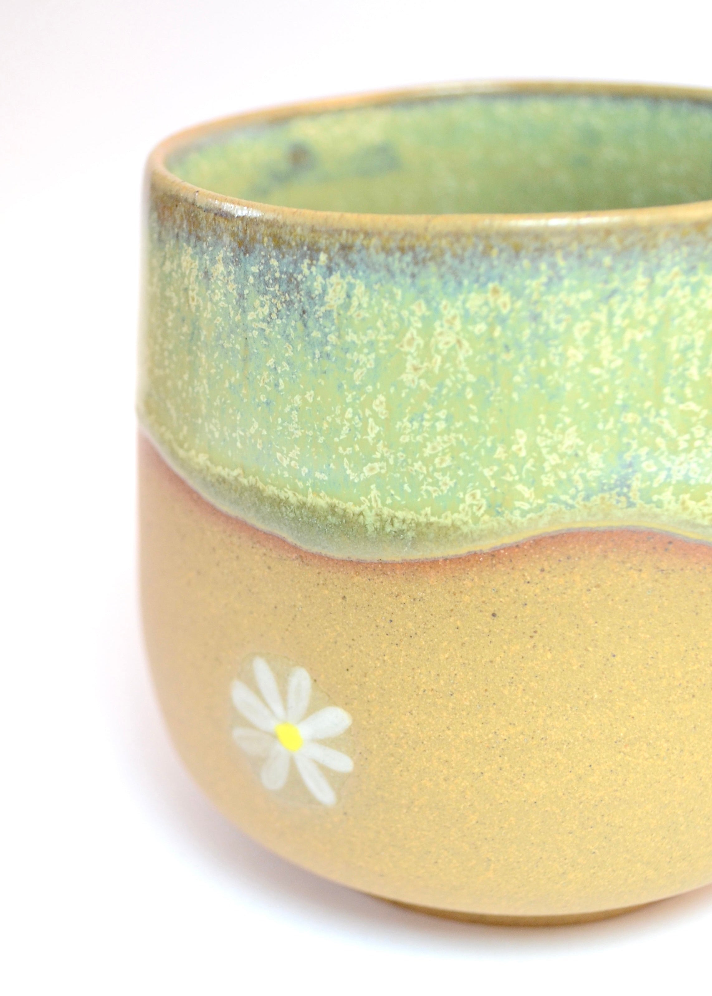 A close-up of a handmade ceramic cup with a two-tone glaze. The top half has a textured light green glaze, and the bottom half is a matte sandy beige with a small white daisy painted on it.